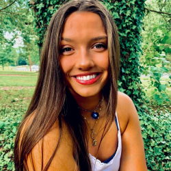 A young woman with long brown hair and a bright smile sits outdoors in front of a tree covered in ivy, wearing a white top and layered necklaces. Green foliage and grass fill the background.