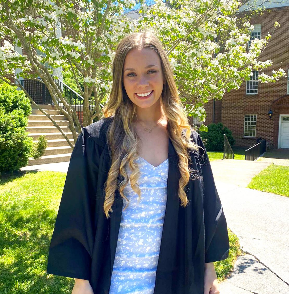 A young woman with long, wavy blonde hair smiles outdoors, wearing a black graduation gown over a light blue dress with white patterns. She stands in front of blooming trees and a brick building on a sunny day.