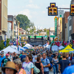A large crowd fills a downtown street lined with tents and flags at a daytime festival in Bristol, Tennessee-Virginia. A sign reading Bristol VA TENN arches over the street near a concert stage.