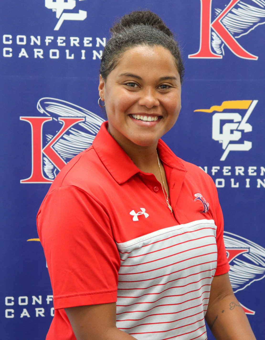 A smiling woman with dark hair in a bun wears a red and white striped polo shirt. She stands in front of a blue backdrop with Conference Carolinas and sports logos.