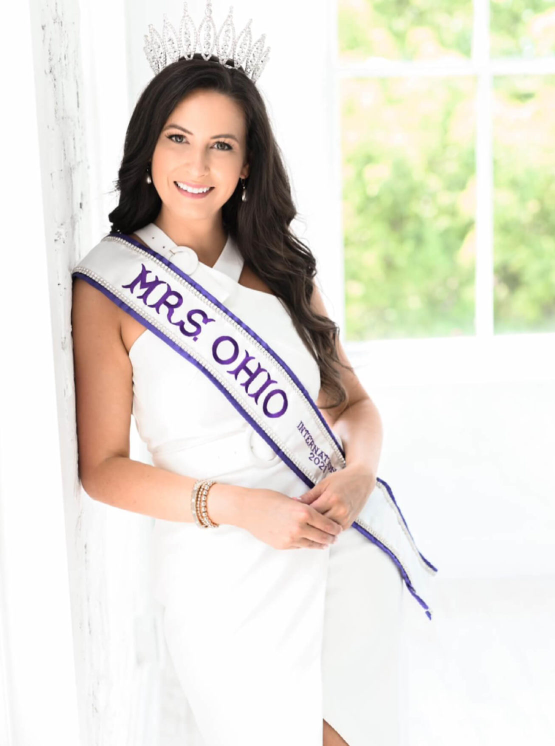 A woman wearing a white dress, a crown, and a sash that reads MRS. OHIO stands smiling by a white wall with a bright window and greenery in the background.