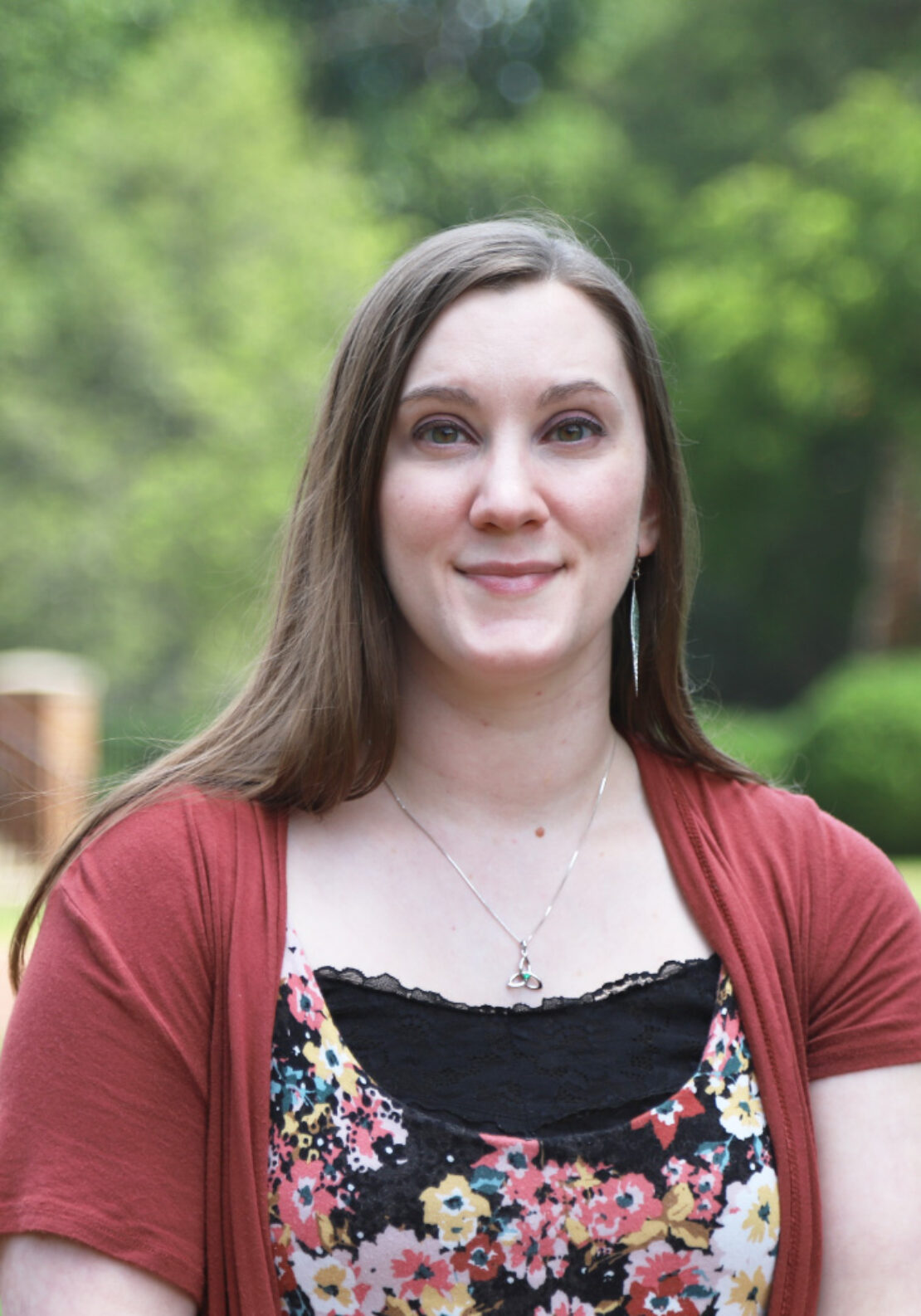 A woman with long brown hair wearing a floral top, a rust-colored cardigan, and a necklace stands outdoors, smiling slightly. The background is blurred greenery.