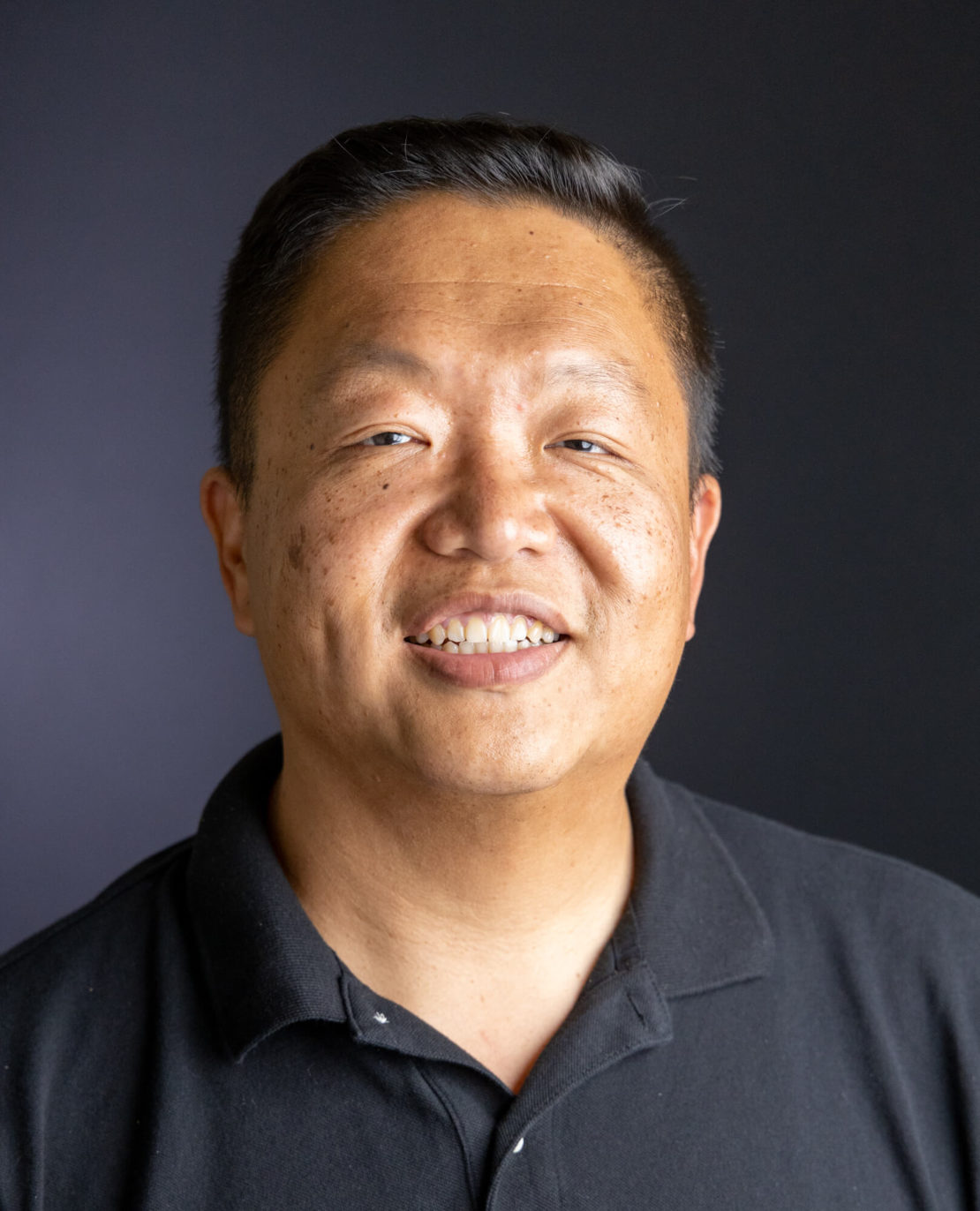 A man with short black hair, wearing a black polo shirt, smiles in front of a dark, plain background.