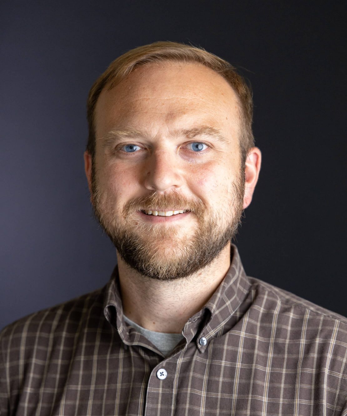 A man with short light brown hair, a beard, and blue eyes, wearing a brown plaid shirt, smiles at the camera against a plain dark background.