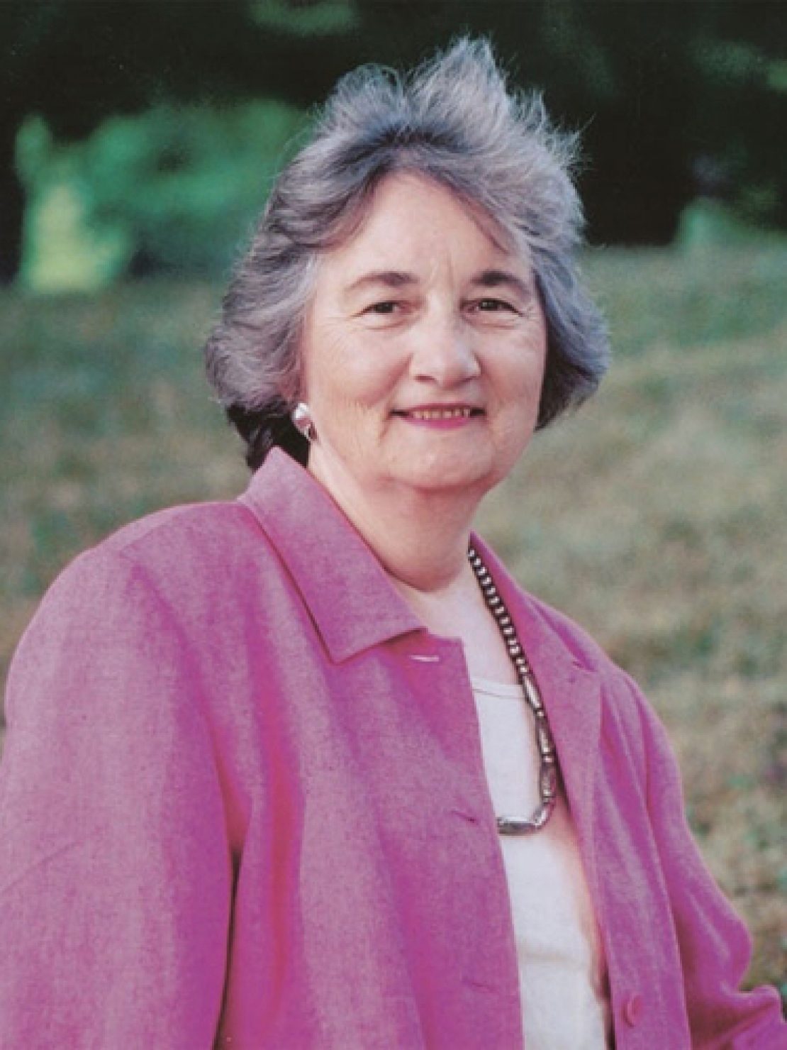 An older woman with short gray hair smiles gently, wearing a light pink jacket over a white top and a long necklace, standing outdoors among grass and trees, embodying the warmth of faith and culture.