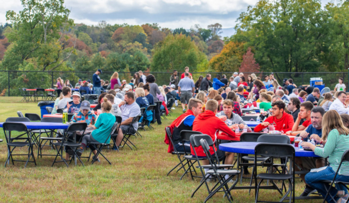 A large group of people sits at round tables outdoors on a grassy field, enjoying food and conversation during a community gathering or picnic on a cloudy day with trees in the background.