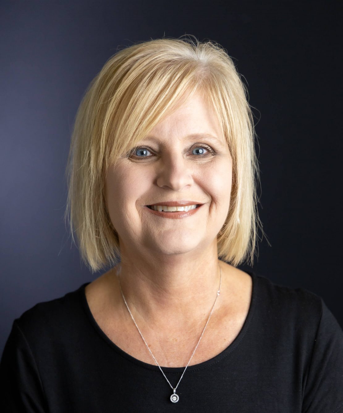 A woman with short blonde hair and blue eyes smiles at the camera. She is wearing a black top and a silver necklace with a small pendant, set against a plain dark background.
