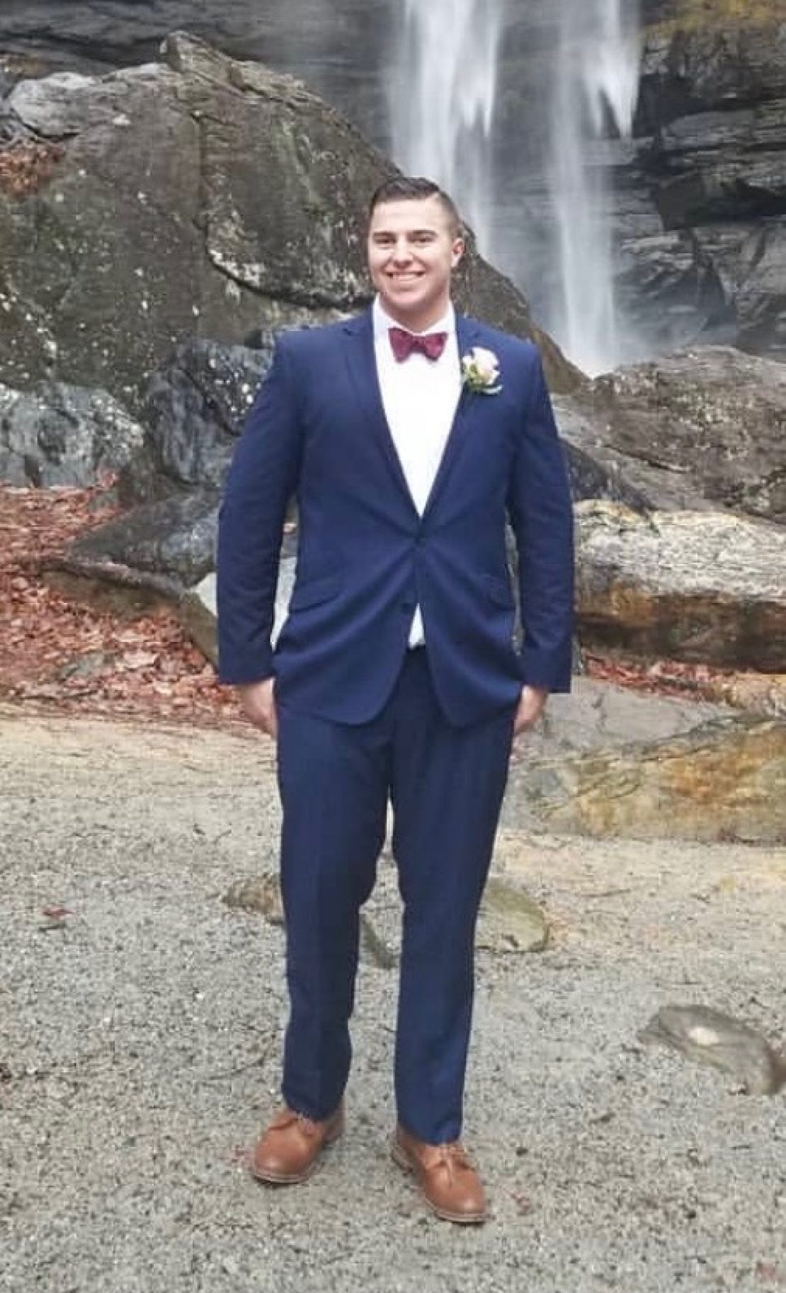 A man wearing a blue suit, white dress shirt, red bow tie, and brown shoes stands outdoors on a gravel path with rocks, fallen leaves, and a waterfall in the background.