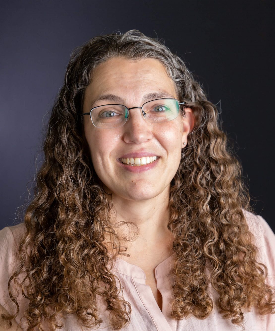 A woman with long, curly brown and gray hair, wearing glasses and a light pink blouse, smiles at the camera against a dark background.