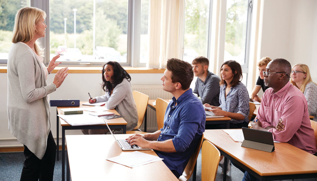 A teacher stands at the front of a bright classroom speaking to a group of adult students seated at desks, some taking notes and using laptops or tablets, all attentively listening.