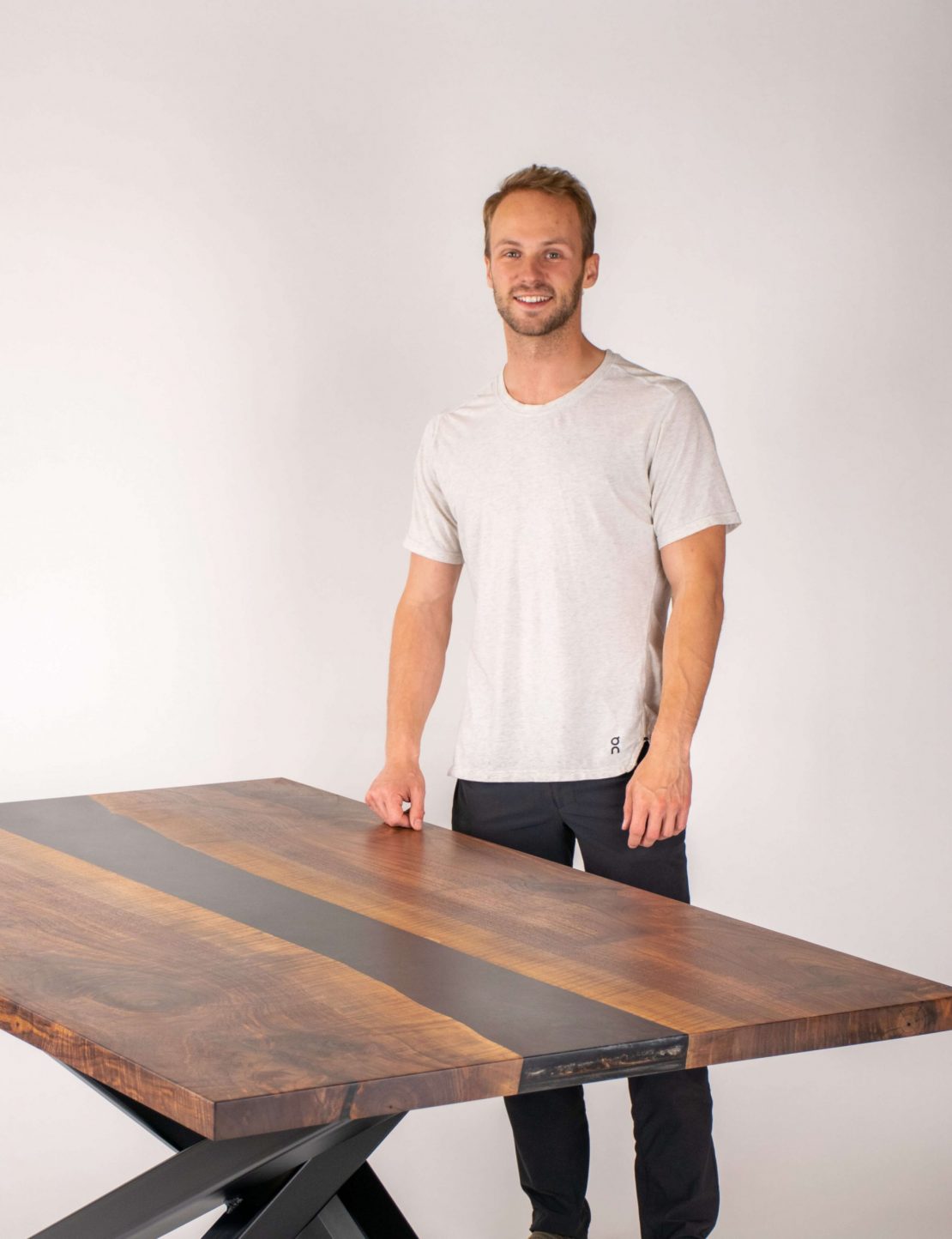 A man in a light gray t-shirt and black pants stands smiling next to a modern wooden table with dark and light wood grain patterns, against a plain white background.
