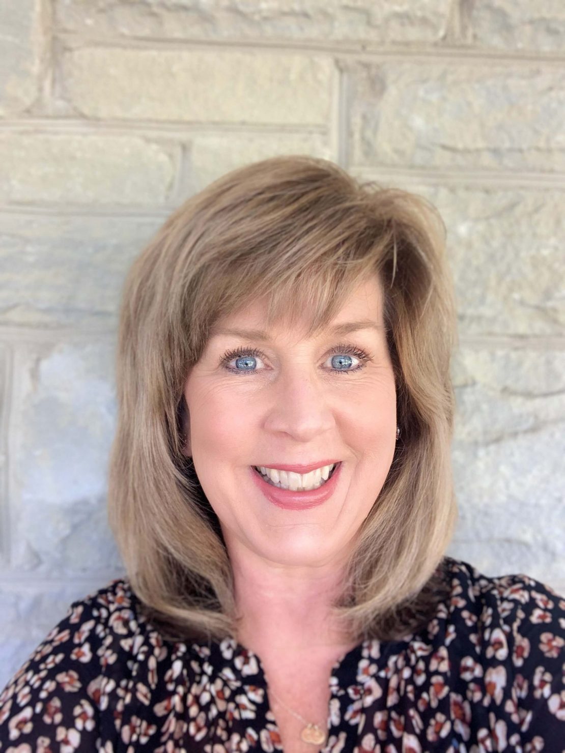 A woman with shoulder-length blonde hair and blue eyes smiles in front of a light gray stone wall. She is wearing a patterned black top and a gold necklace.