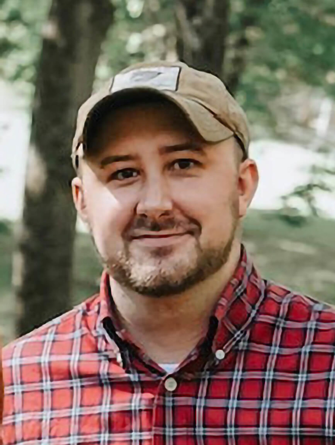 A man with a trimmed beard wearing a tan baseball cap and a red plaid shirt smiles outdoors with trees and greenery in the background.