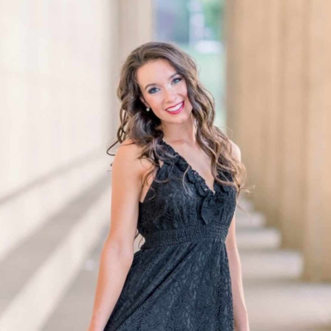 A woman with long brown hair wearing a sleeveless black dress and tan wedge sandals stands outside between tall columns, smiling and holding the sides of her dress.