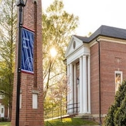 Red brick building with white columns next to a brick clock tower, surrounded by trees and a lamp post. Sunlight filters through the trees, creating a warm and inviting atmosphere.