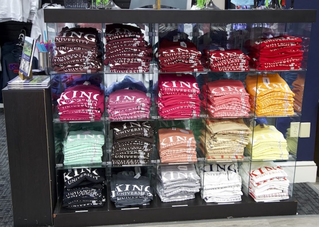 A glass display case holds neatly folded stacks of KING University T-shirts in various colors, arranged by shade in three rows and four columns inside a store.