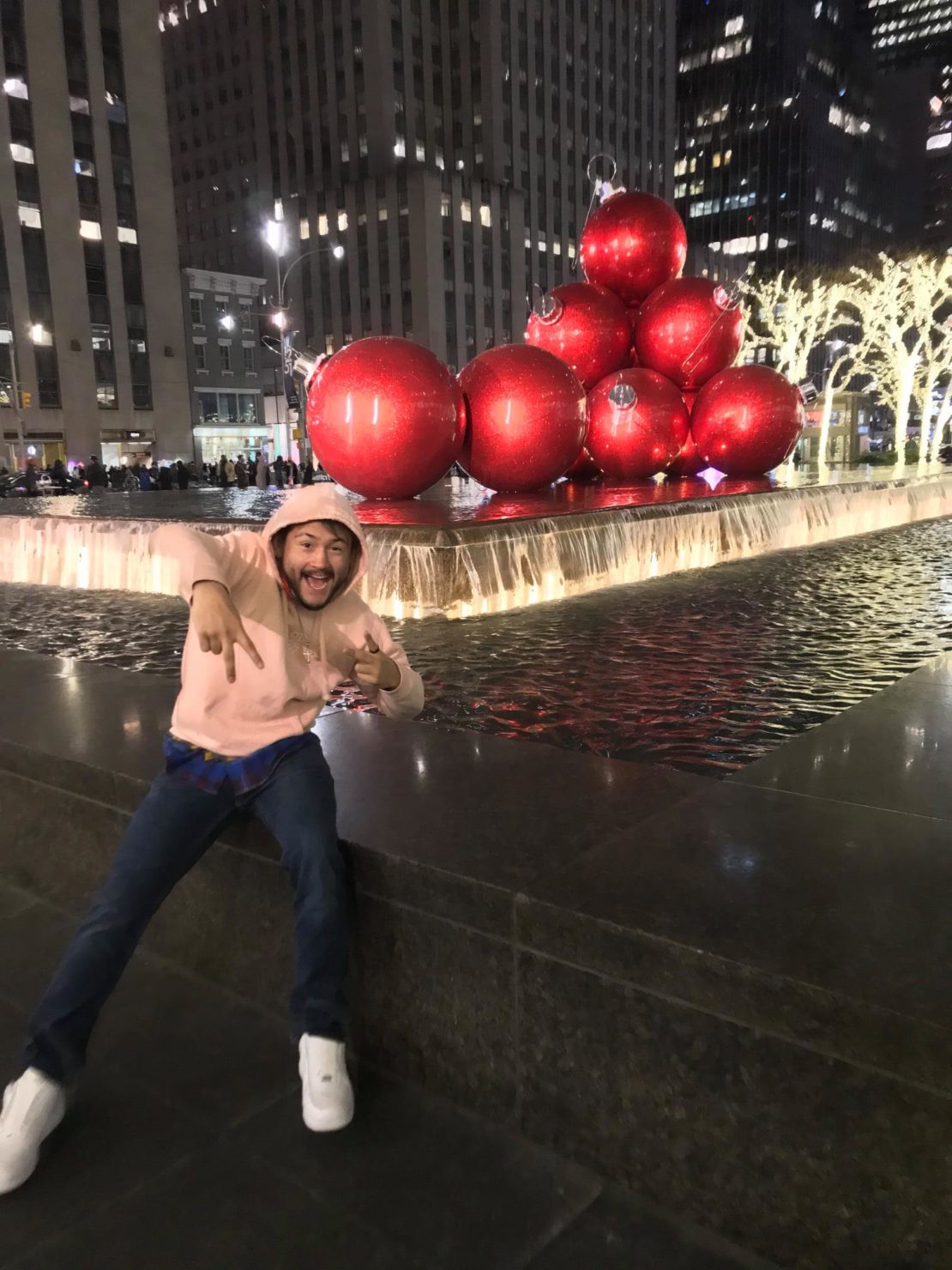 A man in a pink hoodie poses playfully in front of a fountain with large red Christmas ornament decorations at night in a city plaza with lit trees and tall buildings in the background.