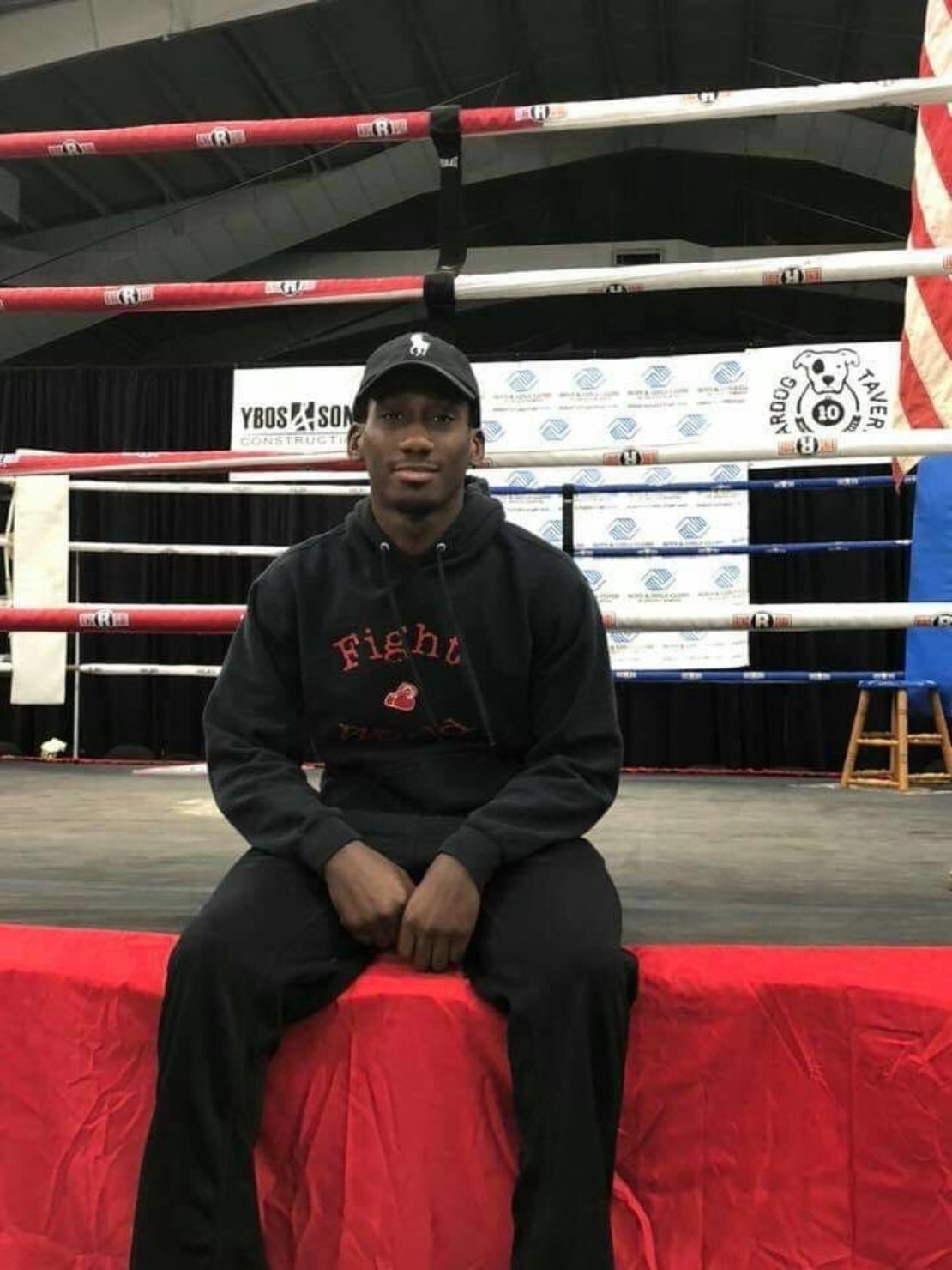 A young man wearing a black cap, black hoodie, and black pants sits on the edge of a red boxing ring, with banners and an American flag visible in the background.