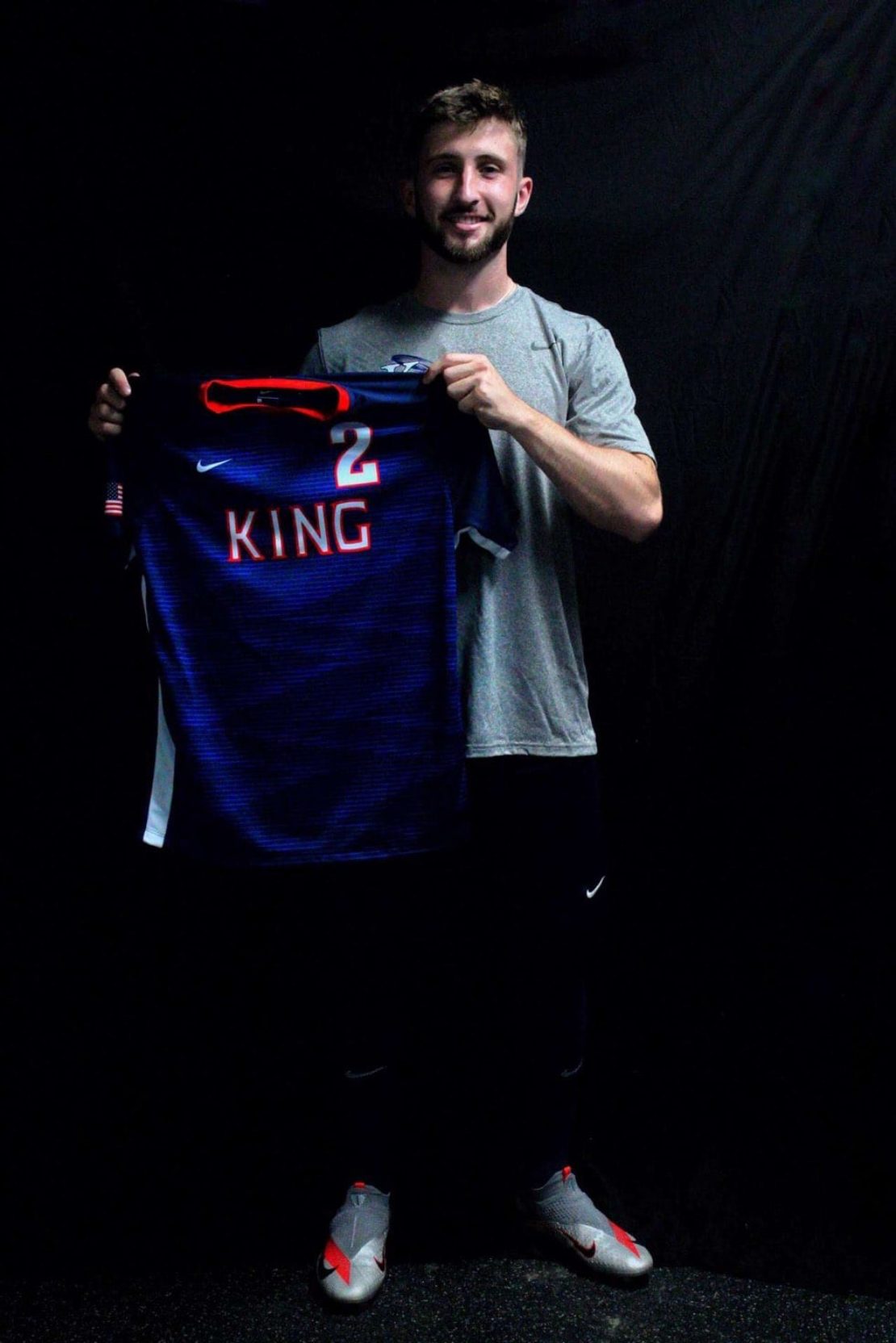 A man in a gray shirt and dark pants stands against a black background, smiling and holding up a dark blue sports jersey with KING and the number 2 printed on the back in white letters.