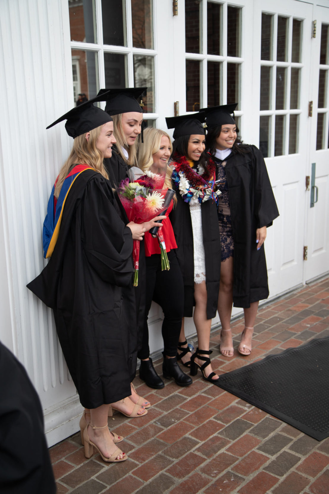 Four graduates in caps and gowns stand outside with a smiling woman, posing for a photo. Two graduates hold bouquets of flowers, and one wears a colorful flower lei. They are standing on a brick patio by white doors.