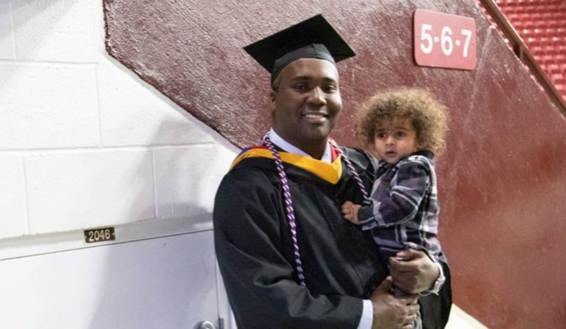 A graduate in a cap and gown smiles while holding a young child with curly hair, standing indoors near a wall with section numbers 5-6-7 visible in the background.