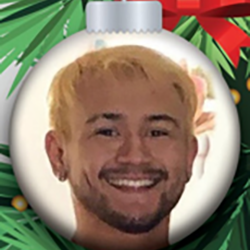 A Christmas tree ornament with a photo of a smiling person with short blond hair and facial hair. The ornament is decorated with green pine needles and a red ribbon.