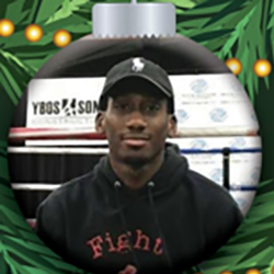 A man wearing a black cap and a black hoodie is pictured inside a Christmas ornament frame with green pine branches and yellow lights. Shelves stacked with boxes appear in the background.