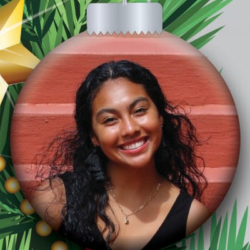 A smiling woman with long curly hair is featured on a Christmas tree ornament. The ornament hangs in front of green pine branches and a gold star decoration.