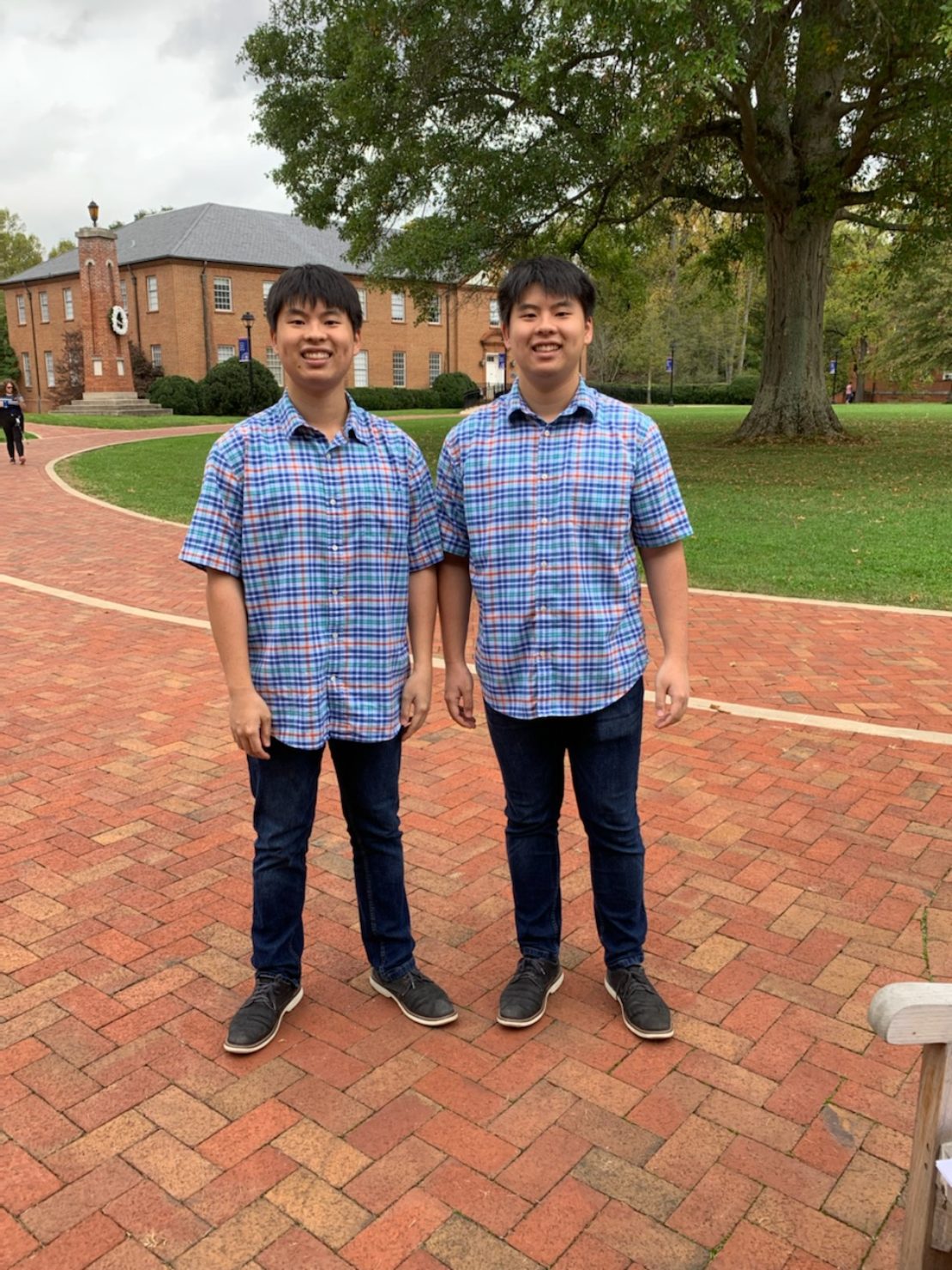 Two young men who appear to be twins stand side by side on a brick walkway outdoors, both wearing matching blue plaid shirts and dark jeans. Trees and a red-brick building are visible in the background.