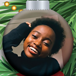 A smiling person with short curly hair and a black top is pictured inside a Christmas tree ornament surrounded by green pine branches.