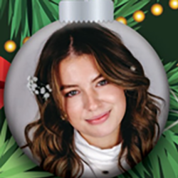A Christmas tree ornament features a portrait of a smiling woman with wavy brown hair, wearing a white top; festive green branches and yellow lights are in the background.