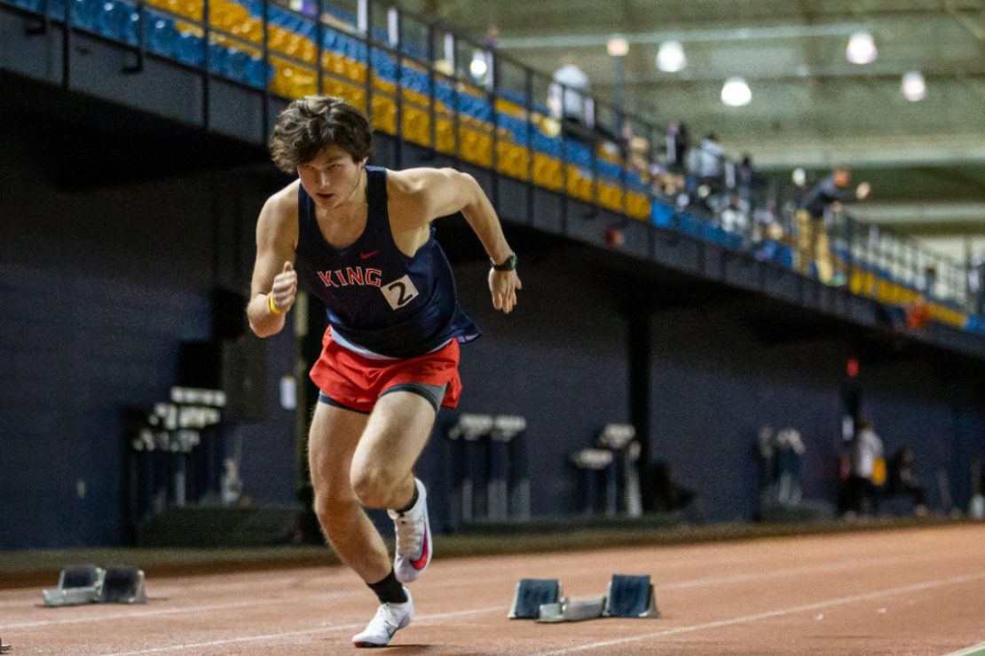 A male athlete wearing a navy tank top and red shorts sprints from the starting blocks on an indoor track, with blurred spectators and empty lanes in the background.