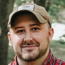 A man with a short beard and mustache wearing a tan baseball cap and a red plaid shirt smiles outdoors with trees and greenery in the background.