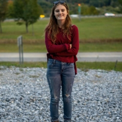 A young woman with long brown hair, wearing a red sweater, blue jeans, and boots, stands on gravel with her arms crossed. There is a grassy field and trees in the background.