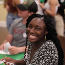 A woman with long braids smiles brightly at the camera while seated at a table, surrounded by other people in a social setting.
