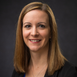 A woman with straight, shoulder-length light brown hair smiles at the camera. She is wearing a black blazer over a blue top and is posed against a dark, plain background.