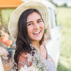 A woman wearing a straw hat smiles outdoors, surrounded by flowers. There is a blurred vehicle and greenery in the background, suggesting a sunny, countryside setting.