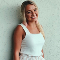 A young woman with long blonde hair, wearing a white sleeveless top and a striped skirt, smiles while standing against a textured light-colored wall.