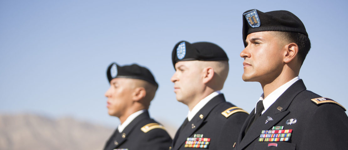 Three military officers in uniform stand in a row outdoors against a clear blue sky, facing forward, with mountains visible in the background.