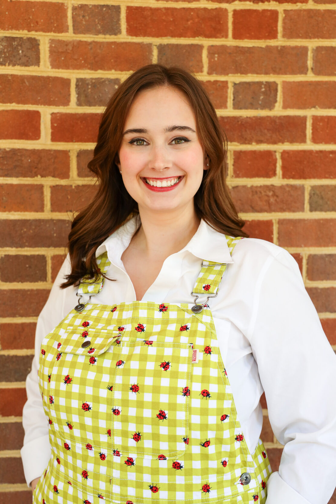 A young woman with long brown hair smiles in front of a brick wall. She wears a white shirt and green plaid overalls with small ladybug designs.