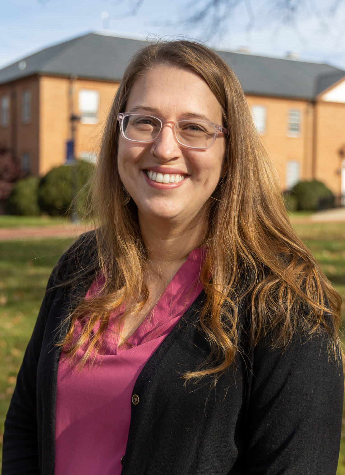 A woman with long light brown hair and glasses smiles while standing outside in front of a brick building, wearing a pink blouse and black cardigan on a sunny day.