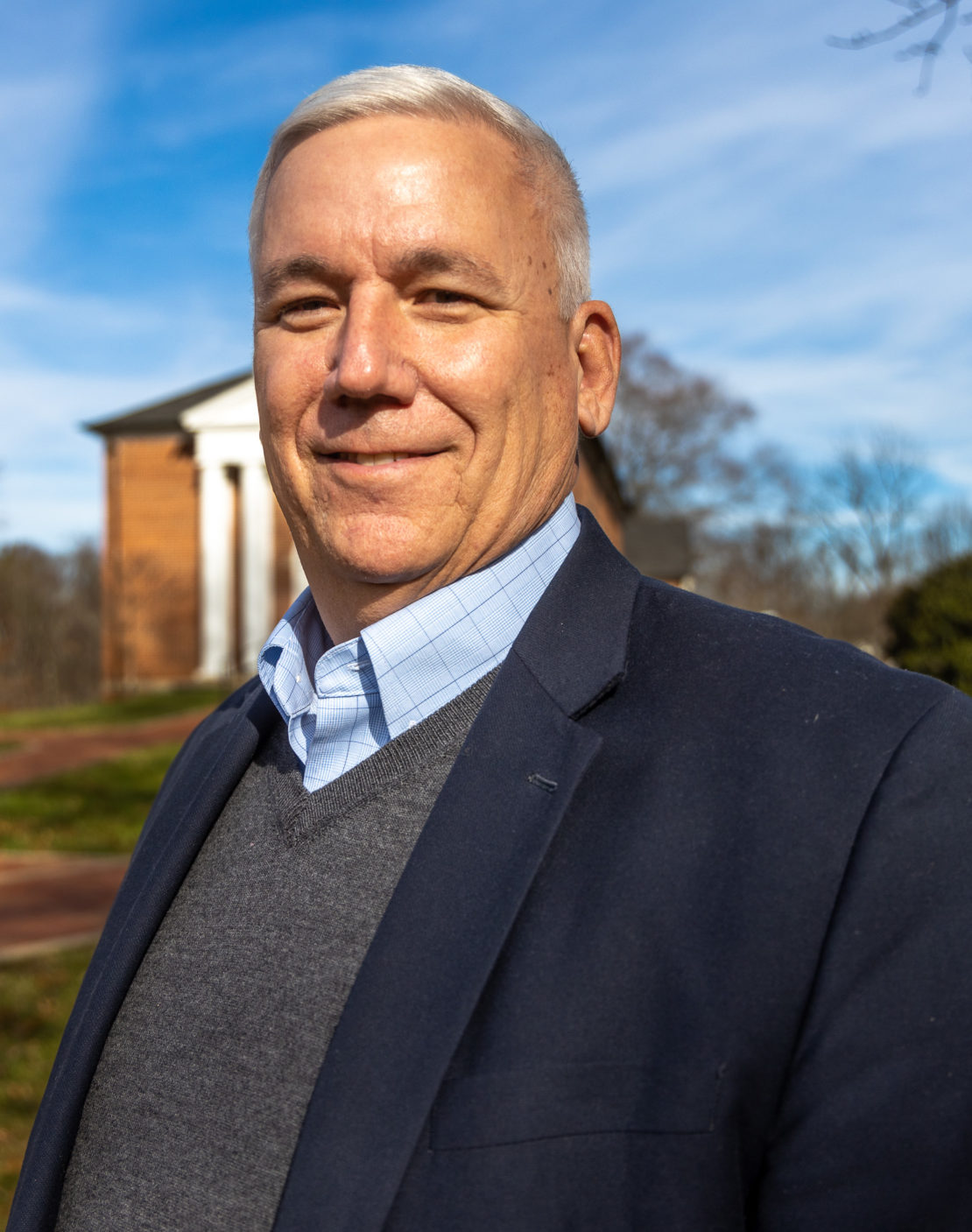 A middle-aged man with short gray hair, wearing a navy blazer, blue dress shirt, and gray sweater, stands outdoors smiling, with a brick building and blue sky in the background.