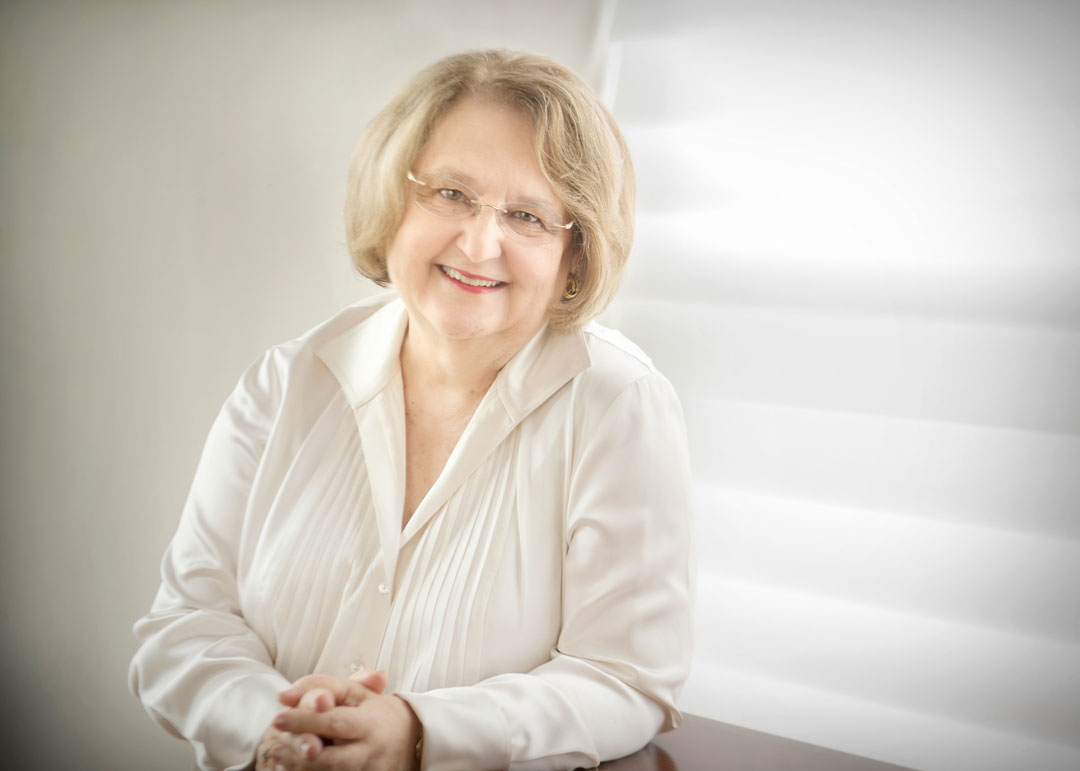 An older woman with short, light hair and glasses, smiling warmly while wearing a white blouse. She is seated with her hands folded, in front of a softly lit, white background with horizontal blinds.