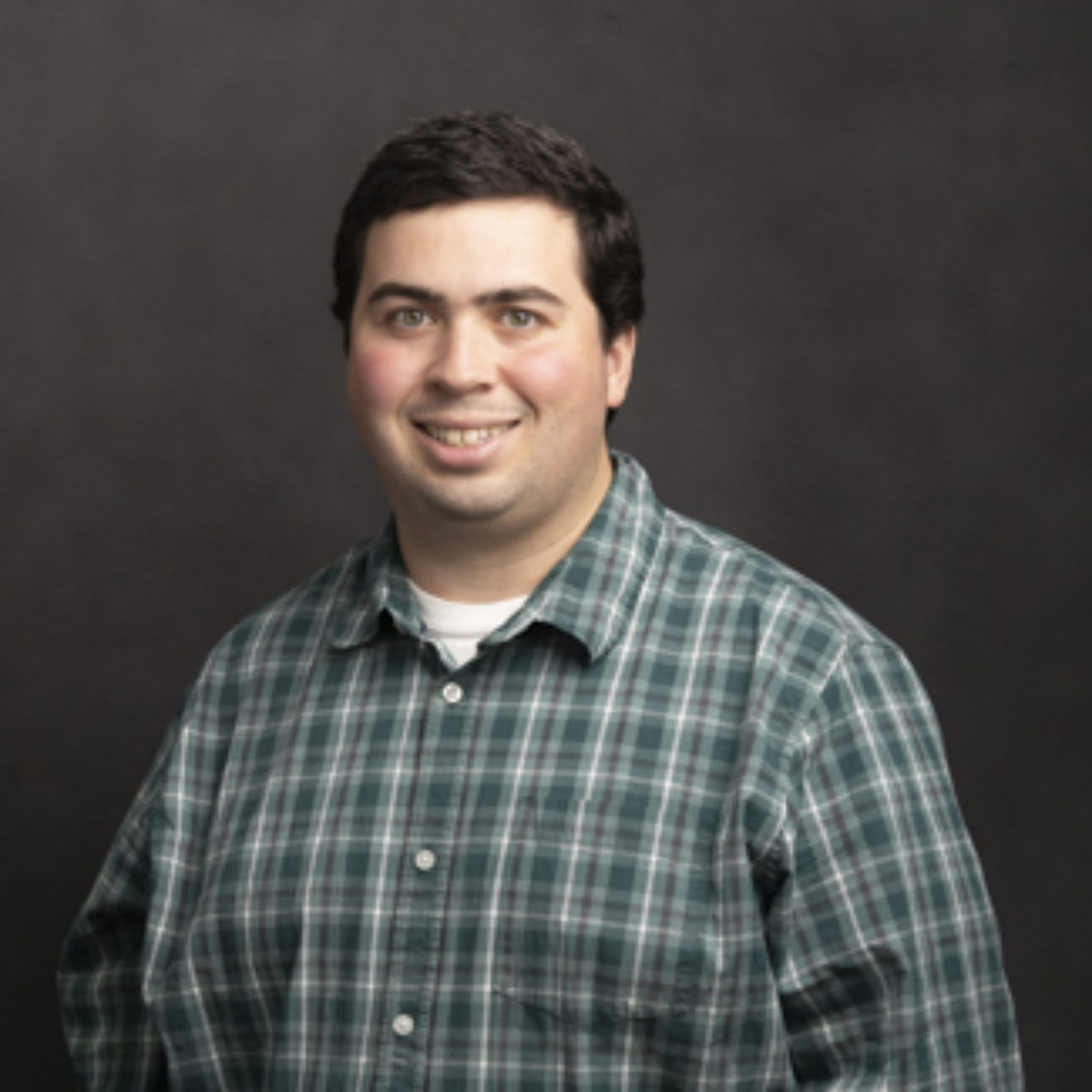 A man with short dark hair smiles at the camera. He is wearing a green and white plaid shirt over a white t-shirt and is standing in front of a dark, plain background.