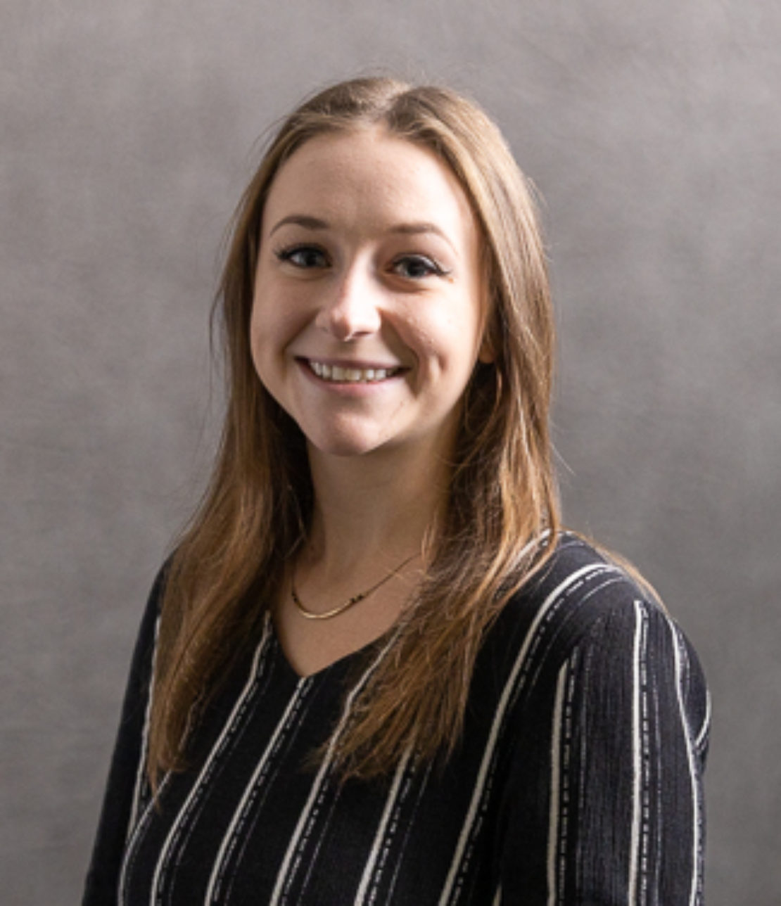 A young woman with straight light brown hair, wearing a black top with white vertical stripes and a necklace, smiles in front of a plain gray background.