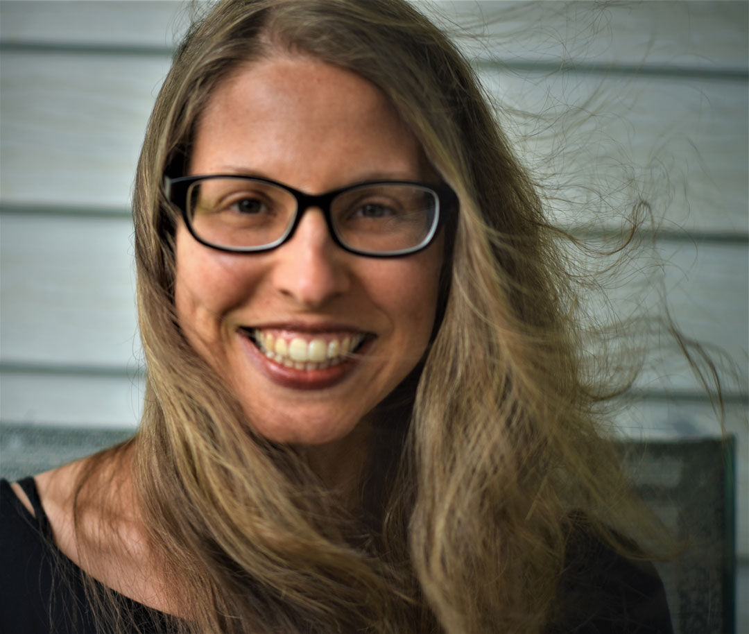 A woman with long, light brown hair and glasses smiles at the camera. She is wearing a black top and sitting in front of a light-colored, wooden panel wall. Her hair is slightly windswept.