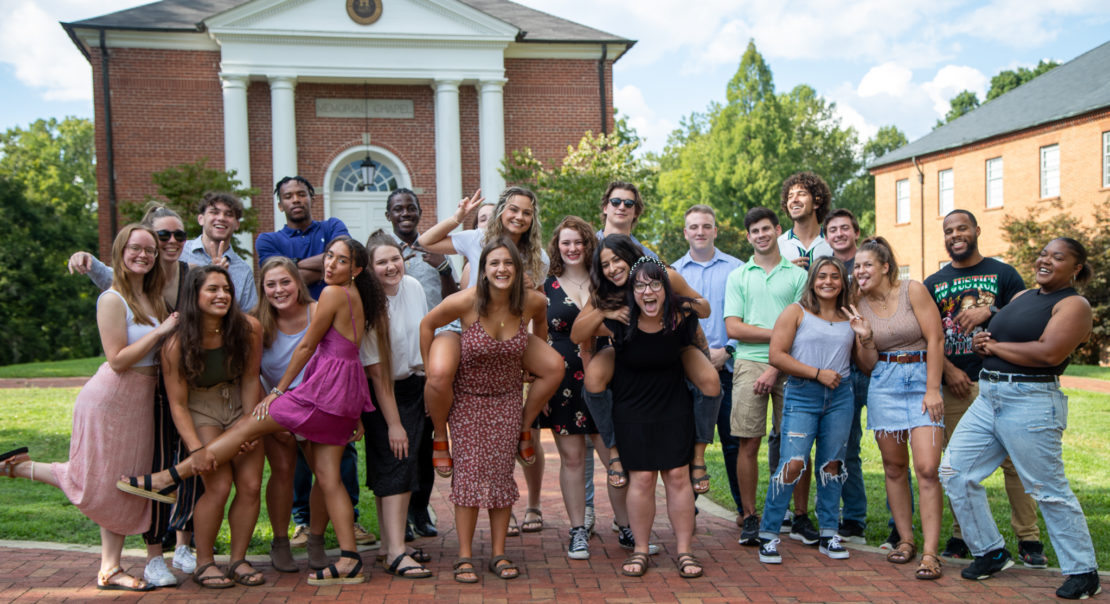 A diverse group of smiling young adults poses together outdoors in front of a brick building, some standing and some giving piggyback rides, enjoying a sunny day.