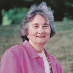 An older woman with gray hair, wearing a pink jacket and a beaded necklace, smiles while standing outdoors with greenery and trees in the background.