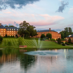 A serene pond with a water fountain in the center, surrounded by green lawns and large red-brick buildings under a pastel sunset sky. Trees and outdoor lights add to the peaceful atmosphere.