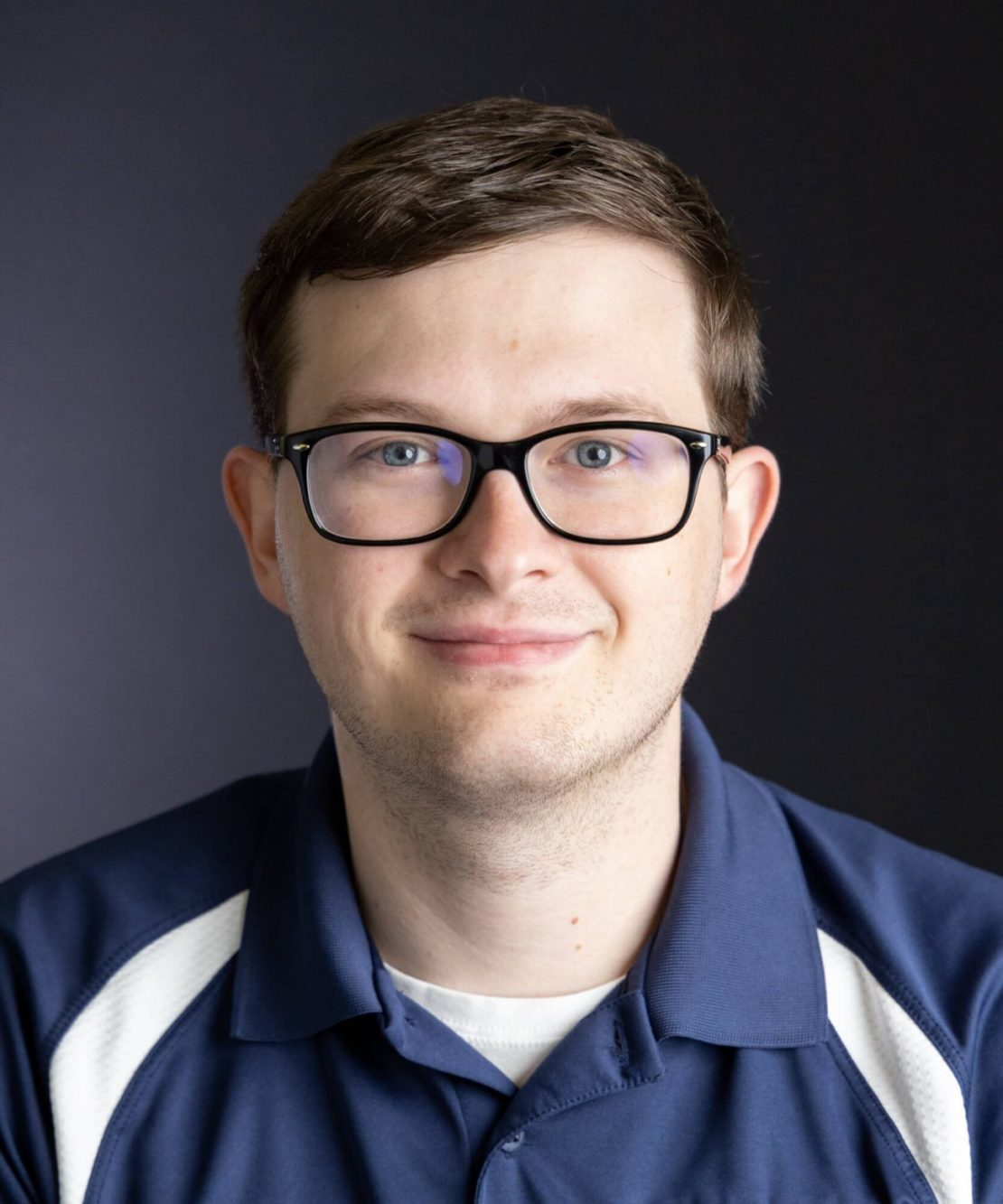 A young man with short brown hair and glasses smiles slightly at the camera. He is wearing a navy blue polo shirt with white accents, and the background is plain and dark.